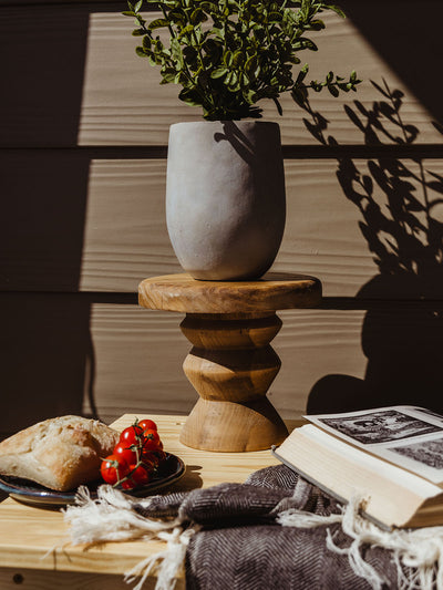 Outdoor flat-lay scene with a rustic wood pedestal riser holding a matte clay vase with greenery, surrounded by an open book, herringbone throw, cherry tomatoes, and crusty bread.
