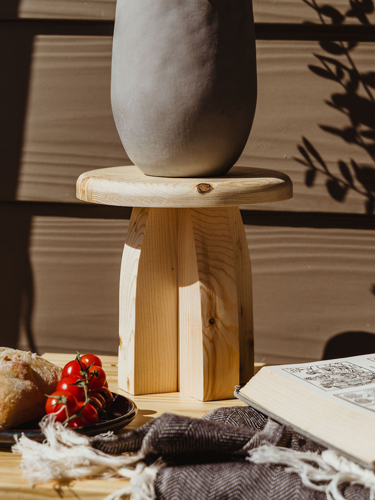 Close-up of a minimalist plant in a smooth gray pot placed on a light wood pedestal, with fresh tomatoes, bread, and an open illustrated book in the foreground.
