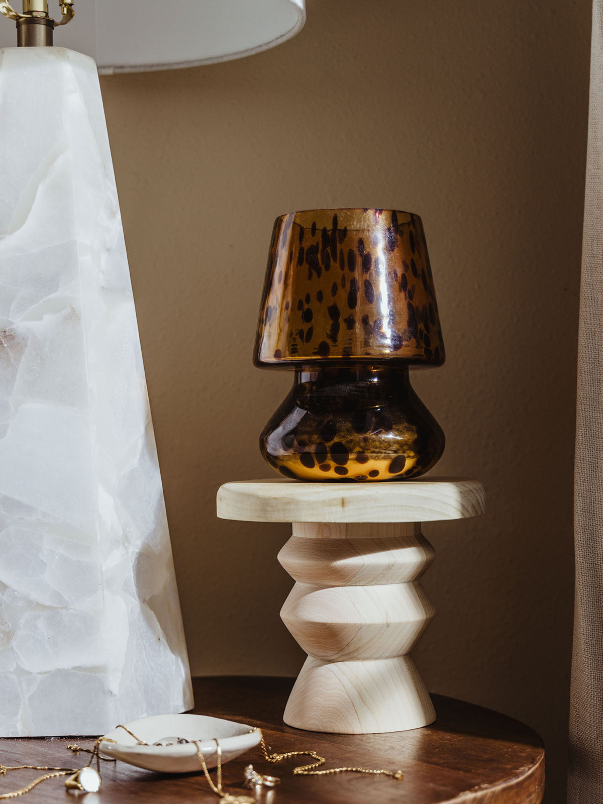 Curved wood pedestal riser styled on a nightstand, holding a tortoiseshell glass lamp next to a white marble lamp and a jewelry dish with gold accessories.