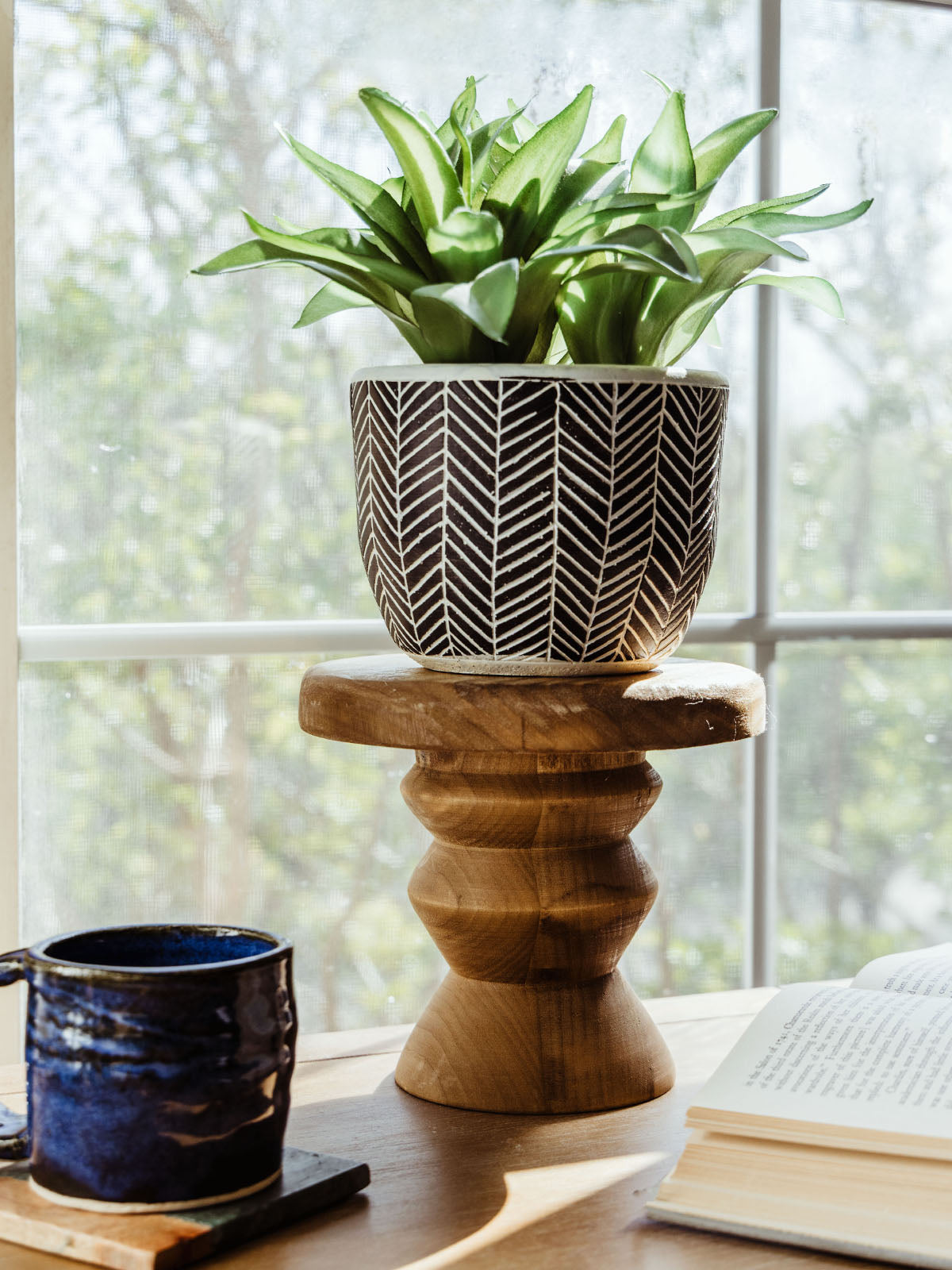 Natural wood pedestal riser holding a black-and-white chevron planter with a green plant, styled on a windowsill beside a ceramic mug and open book.