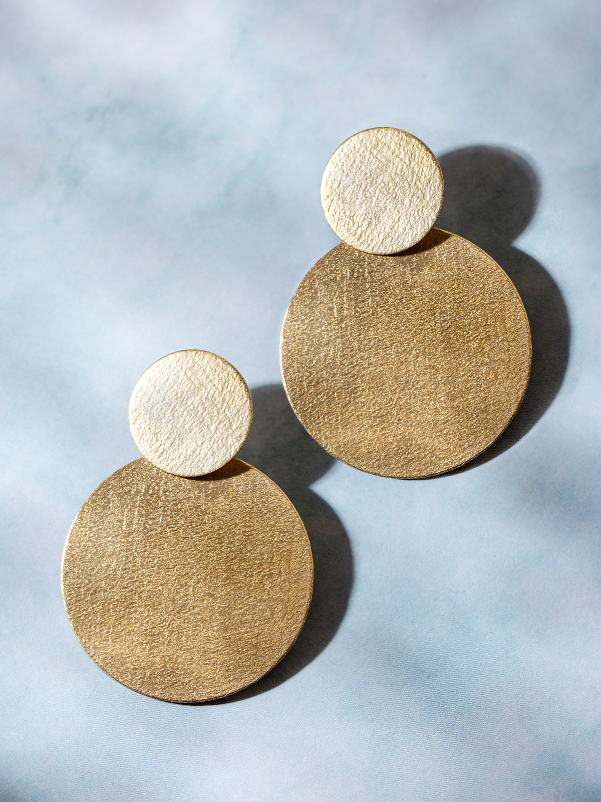 A pair of textured gold statement earrings featuring a double-disc design, displayed on a soft blue background. The brushed gold surface catches the light, highlighting the contemporary aesthetic.