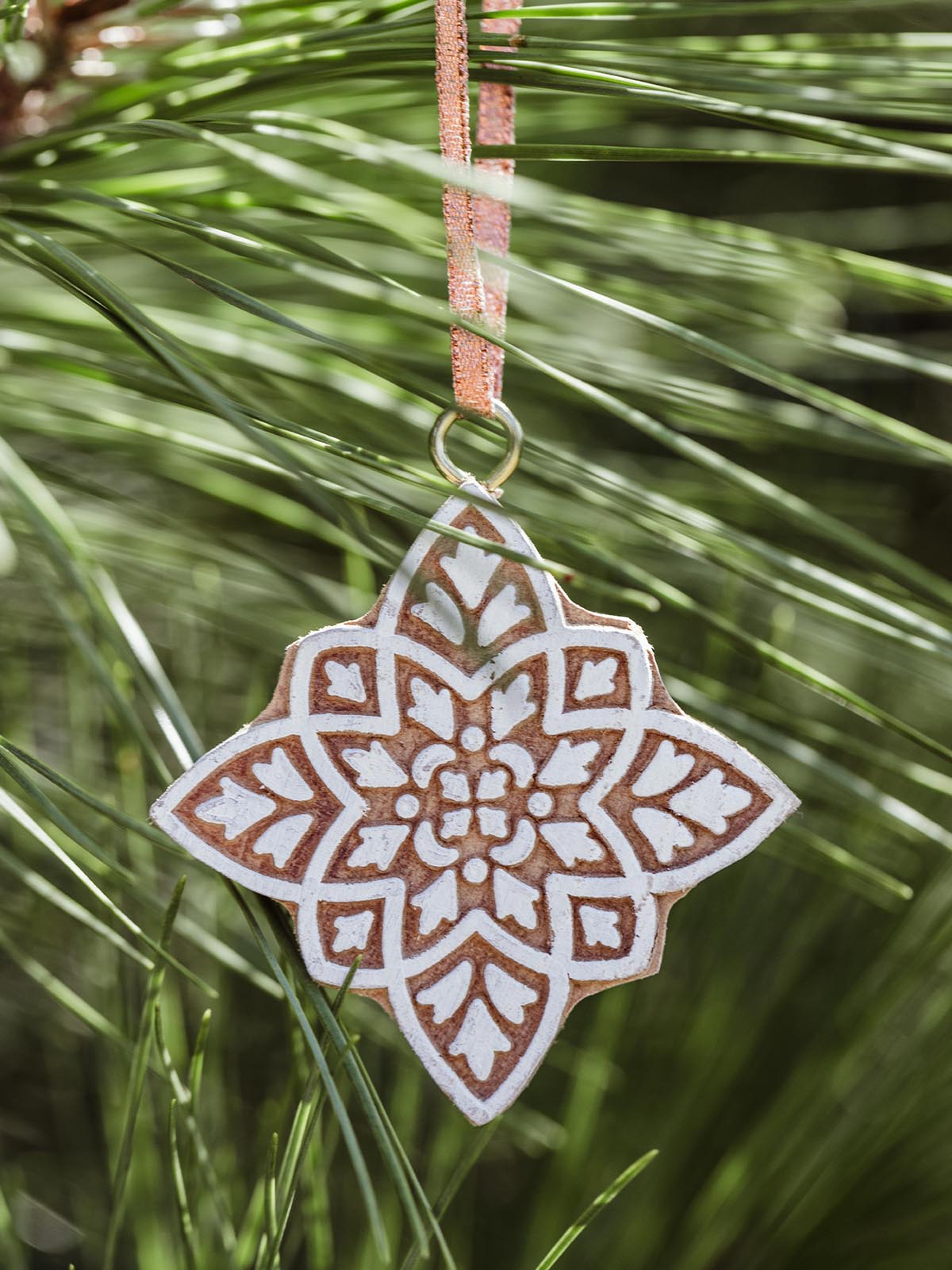 Handcrafted ceramic star-shaped ornament with an intricate white and terracotta pattern, hanging from a rustic ribbon against a backdrop of green pine needles.