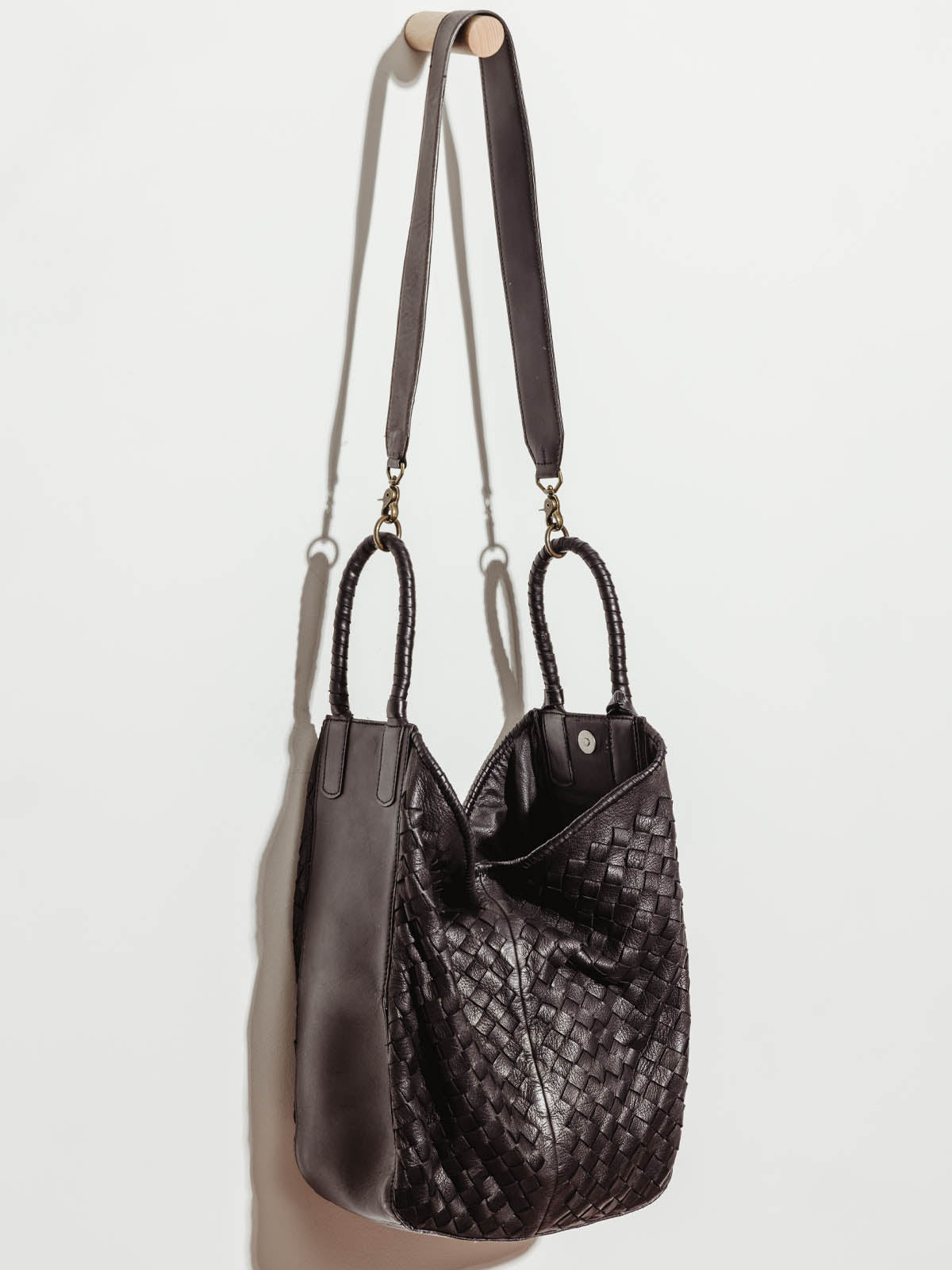 Black leather all day tote unclasped, hanging by shoulder strap on white wall.