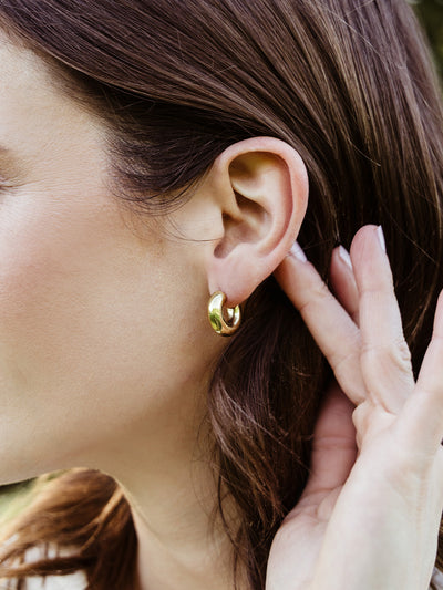 Side profile of a woman wearing medium-sized gold hoop earrings, catching the natural light. Her hand is raised to her hair, showcasing elegant, timeless jewelry perfect for everyday wear.