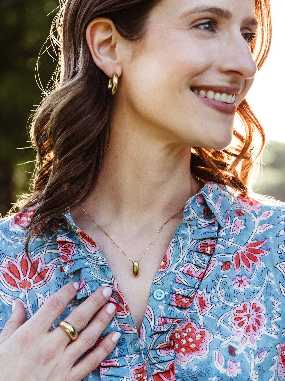 Woman outdoors smiling in the sunlight, wearing gold hoop earrings, a gold pendant necklace, and a chunky gold ring. She pairs the jewelry with a floral blue and red blouse, highlighting an effortlessly chic look.