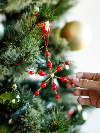 Handcrafted red and gold beaded snowflake ornament made from recycled paper beads, hanging on a Christmas tree branch.