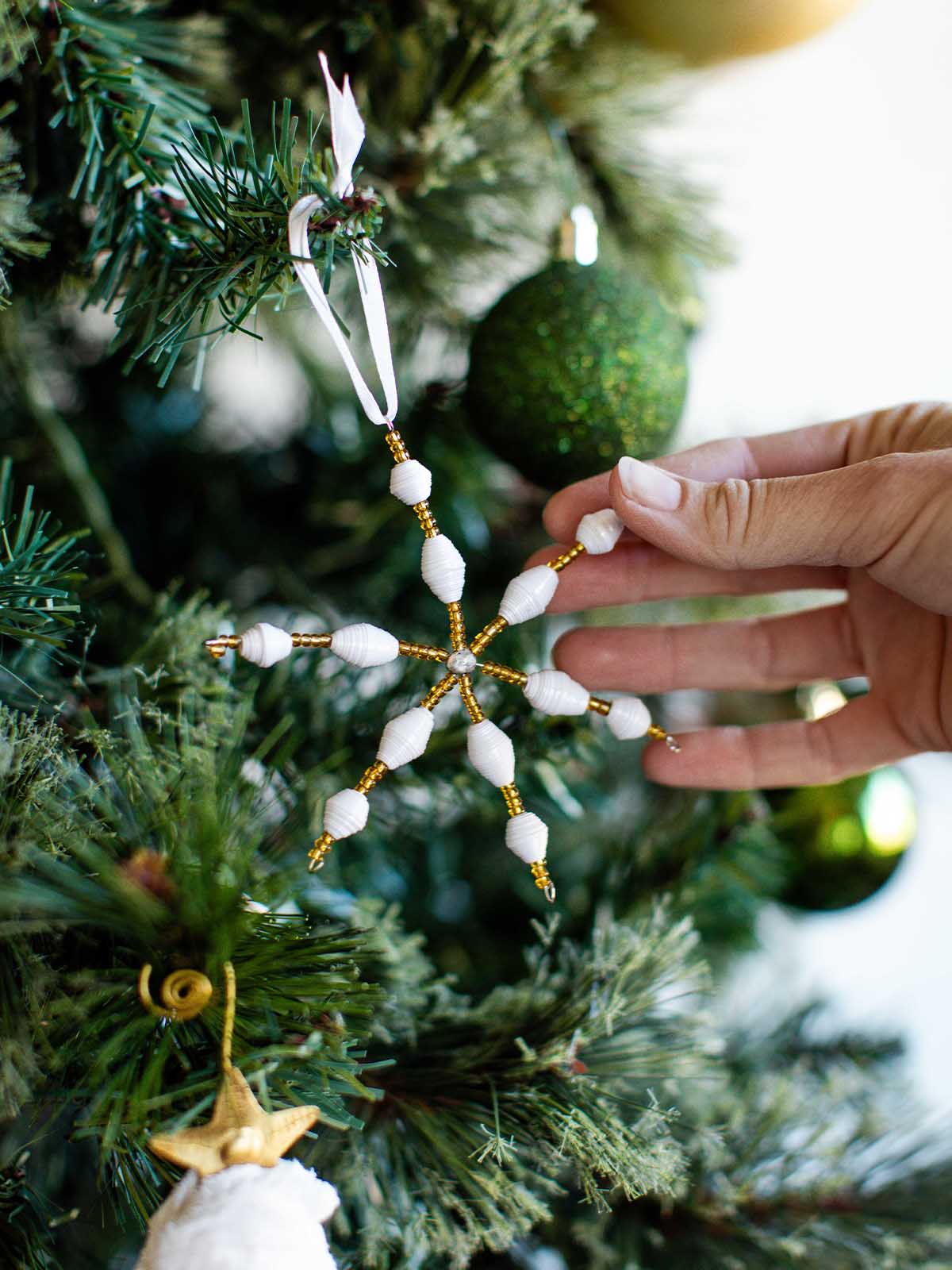 White and gold handcrafted paper bead snowflake ornament with ribbon hanger, displayed on a Christmas tree.