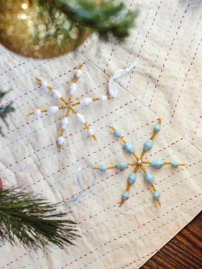 Assorted handcrafted paper bead snowflake ornaments in white and light blue tones resting on a quilted fabric beneath a Christmas tree.