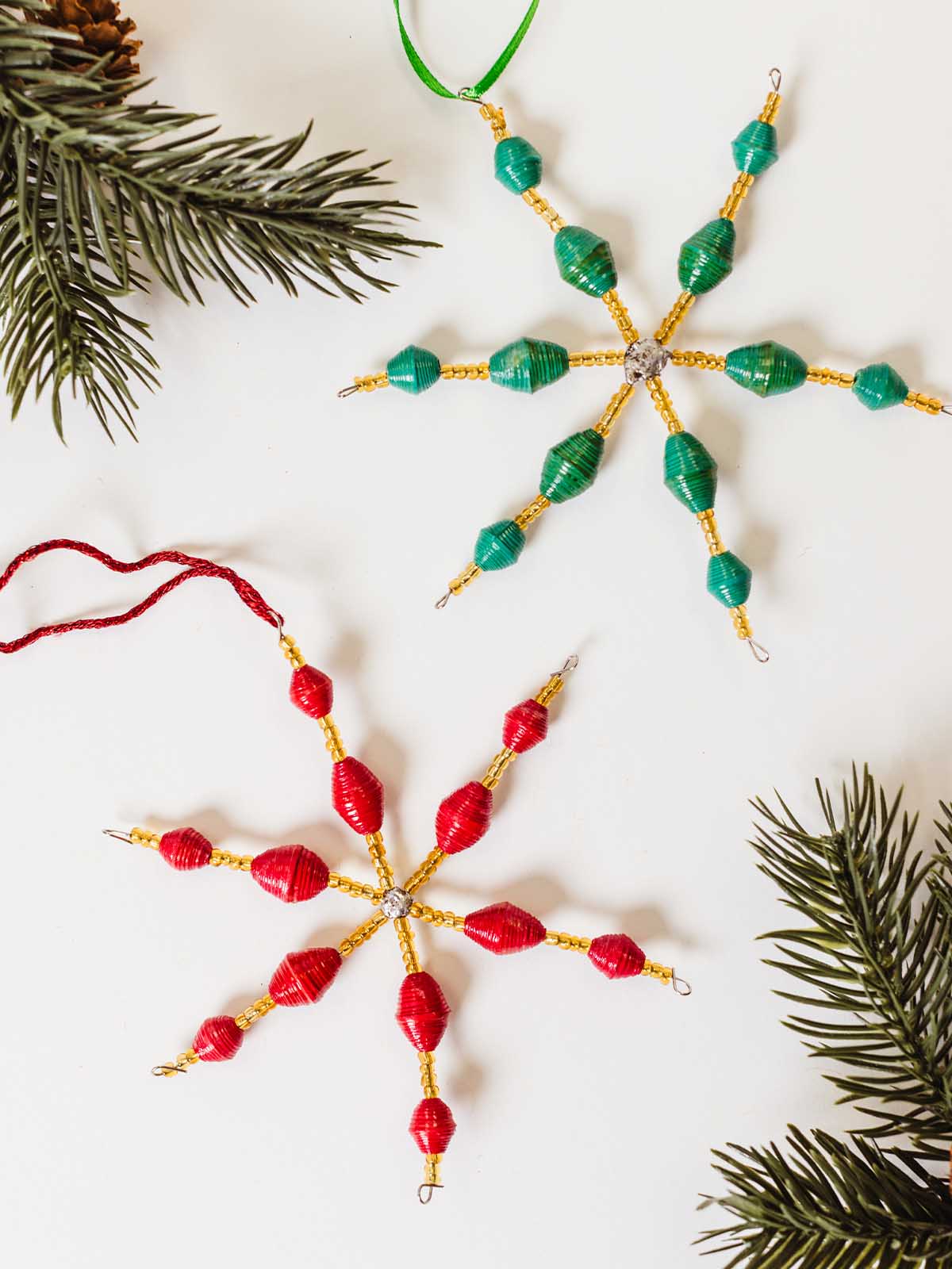 Red and green paper bead snowflake ornaments with gold accents and ribbon loops, styled with evergreen branches on a white background.