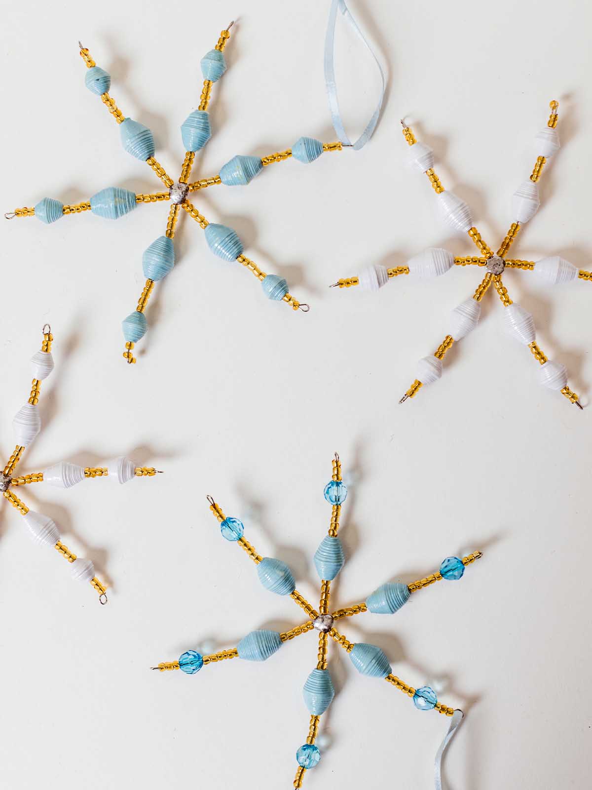 Light blue and white paper bead snowflake ornaments with gold bead accents, arranged on a white background for holiday décor.