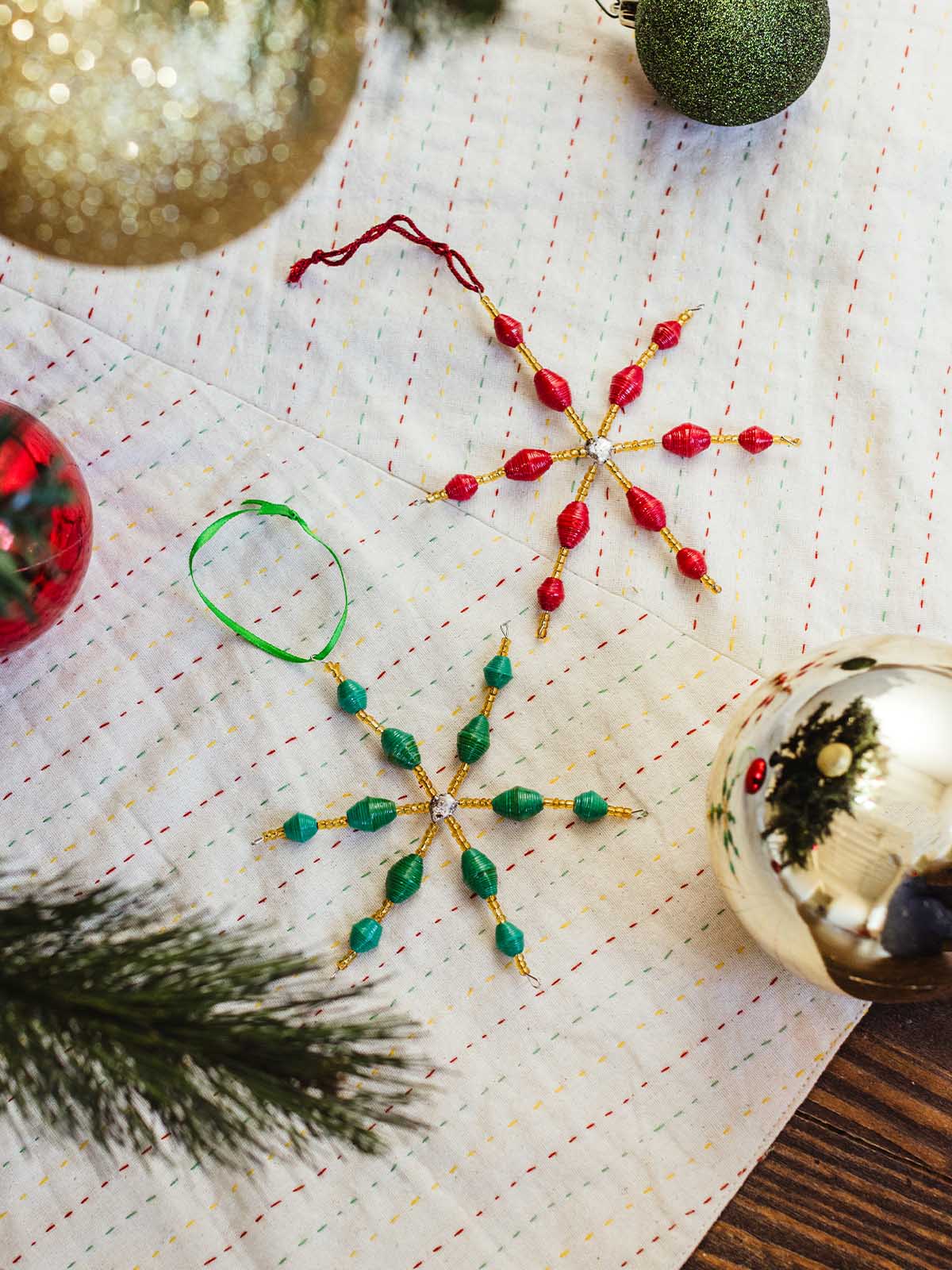 Red and green handcrafted paper bead snowflake ornaments with gold accents resting on a stitched holiday tree skirt surrounded by Christmas ornaments and pine branches.