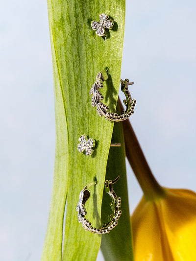 Pair of silver earrings displayed on a fresh green leaf, with a yellow tulip in the background. The earring set includes two crystal-studded flower studs and a pair of dainty silver hoops with beaded accents, creating a nature-inspired aesthetic.