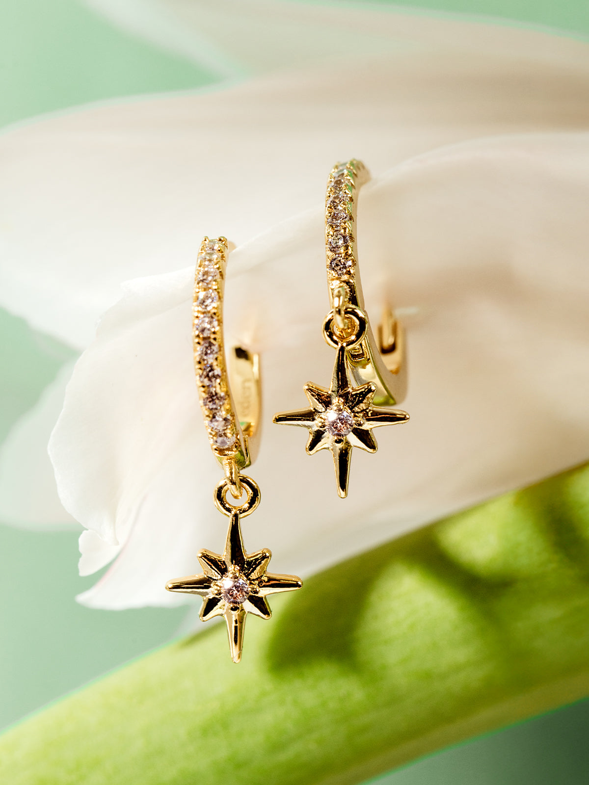 Pair of gold hoop earrings with sparkling pavé stones and dangling eight-point star charms, styled on a white flower petal with a mint green background.