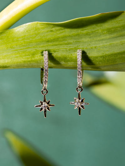 Pair of silver hoop earrings with pavé stones and dangling star charms, hanging delicately from a green leaf against a teal background.