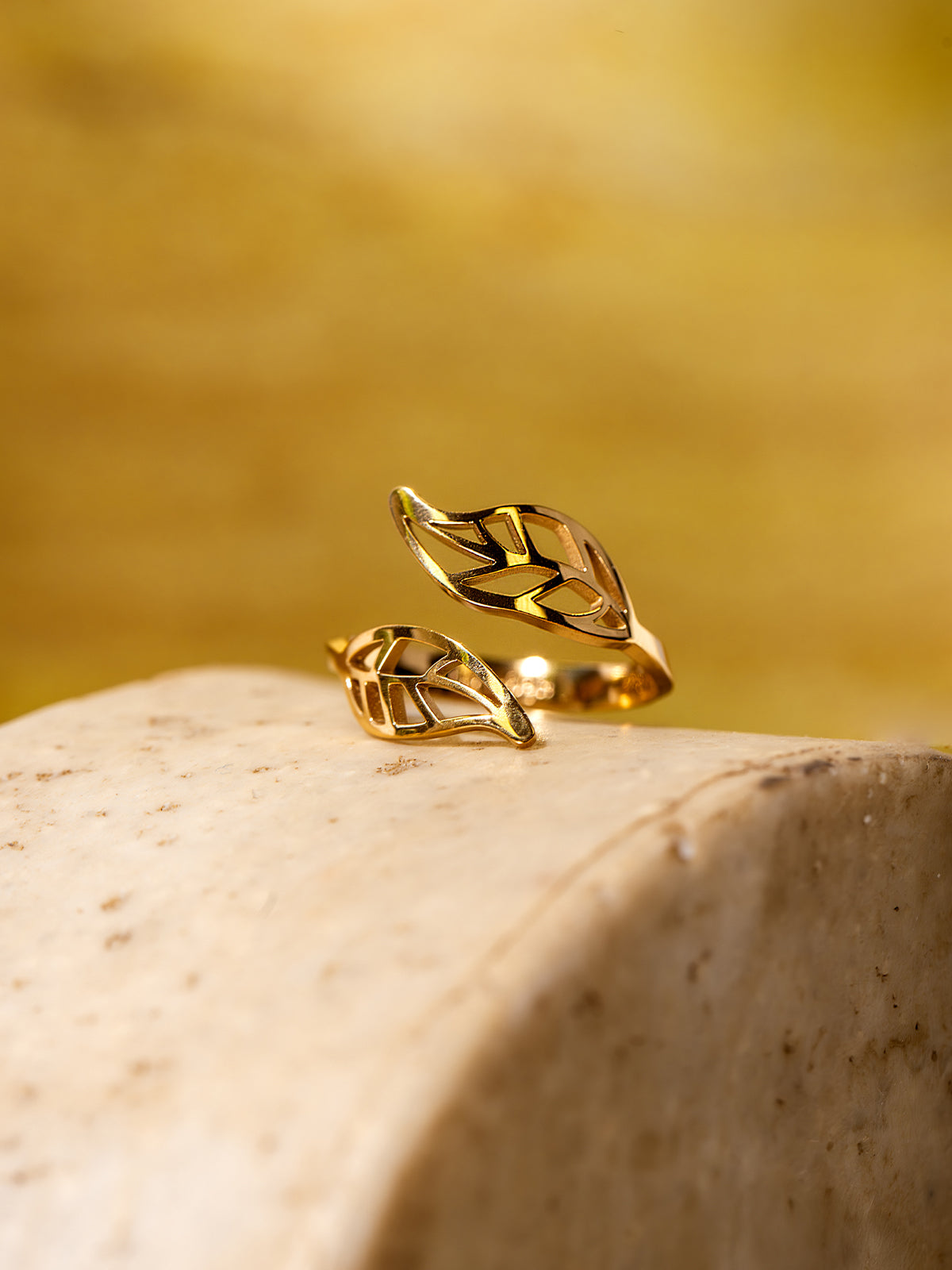 Gold wrap ring with two openwork leaf shapes, displayed on a smooth beige stone surface with a warm yellow background.