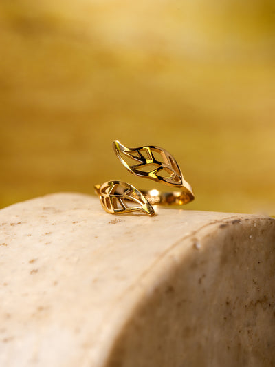 Gold wrap ring with two openwork leaf shapes, displayed on a smooth beige stone surface with a warm yellow background.