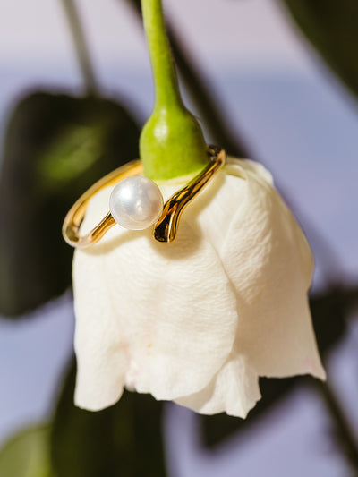 A gold ring with a single pearl elegantly placed on a white rose bud. The organic curve of the band complements the soft glow of the pearl, creating a romantic and nature-inspired jewelry display.