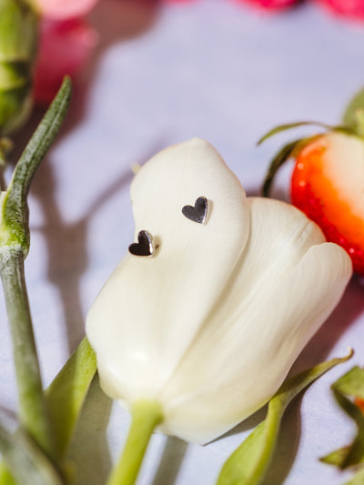 Pair of small silver heart stud earrings resting on a white tulip petal, surrounded by floral accents and a sliced strawberry.