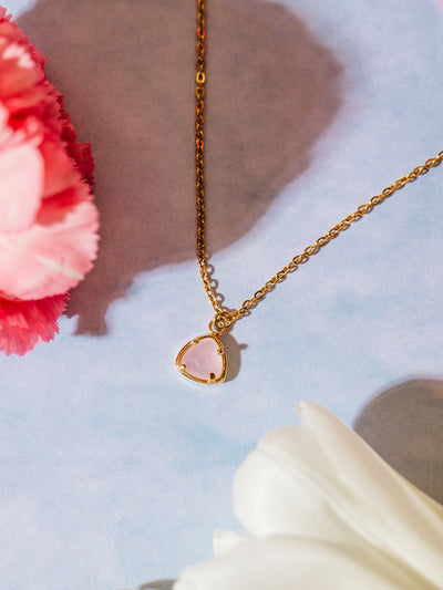 A gold chain necklace with a soft pink faceted gemstone pendant, artfully placed among pink carnations and white tulips. The dreamy composition highlights the romantic and dainty nature of the jewelry.