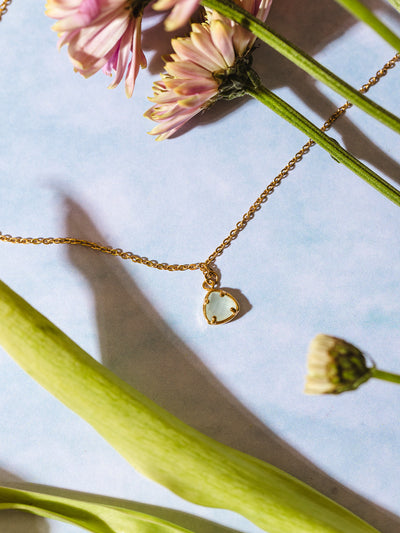 A gold pendant necklace featuring a pale green faceted gemstone, nestled among fresh greenery and pastel florals. The light blue background adds a soft and ethereal touch to the composition.