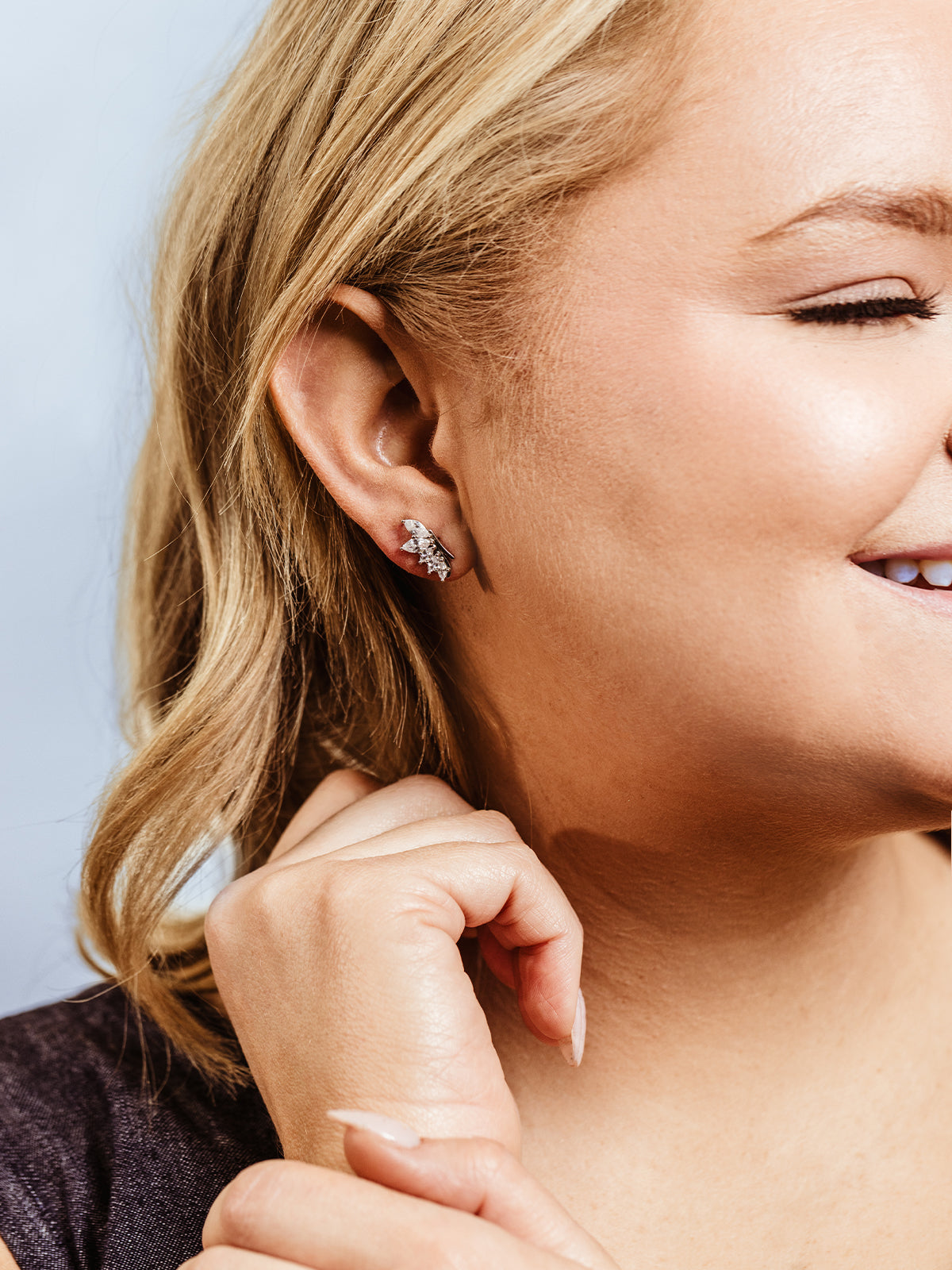 Close-up of a woman with blonde hair smiling while wearing silver leaf-shaped stud earrings with sparkling crystal accents. She gently touches her chin, highlighting the delicate and elegant design of the jewelry.
