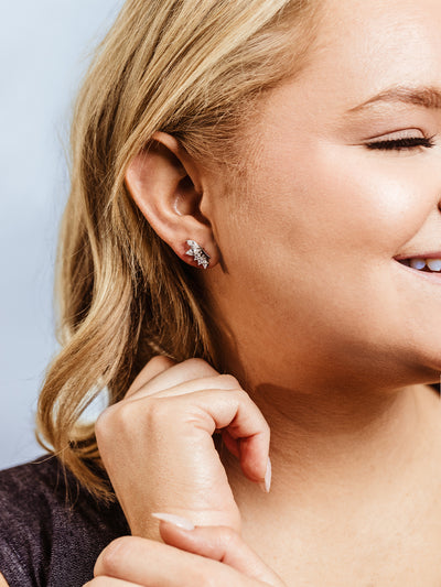Close-up of a woman with blonde hair smiling while wearing silver leaf-shaped stud earrings with sparkling crystal accents. She gently touches her chin, highlighting the delicate and elegant design of the jewelry.