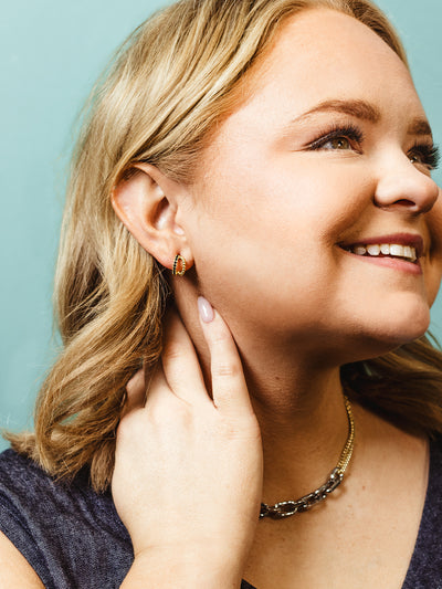 A blonde woman smiles while touching her neck, wearing small gold hoop earrings with intricate beaded details. She pairs them with a bold two-tone chain necklace, adding a stylish contrast to her look against a teal background.