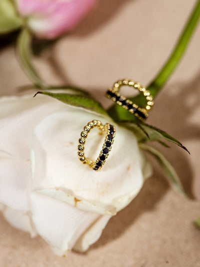 A pair of gold beaded hoop earrings with black gemstone accents elegantly displayed on a soft white rose. The intricate details of the beading and dark stones create a striking, romantic aesthetic