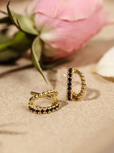 A pair of gold hoop earrings featuring black gemstone inlays and a beaded texture, artistically placed on a neutral sand-toned background. A soft pink rose adds a delicate floral contrast.