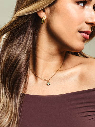 A woman with long, wavy hair wears a delicate gold chain necklace featuring a faceted gemstone pendant in a soft pastel hue. She pairs it with sleek gold drop earrings and an off-the-shoulder top, creating an elegant and modern look.