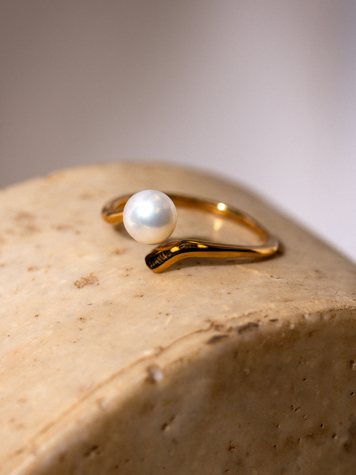 Close-up of a delicate gold ring featuring a single round white pearl set on a curved band, displayed on a textured beige stone surface with soft lighting.