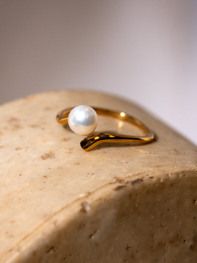 Close-up of a delicate gold ring featuring a single round white pearl set on a curved band, displayed on a textured beige stone surface with soft lighting.