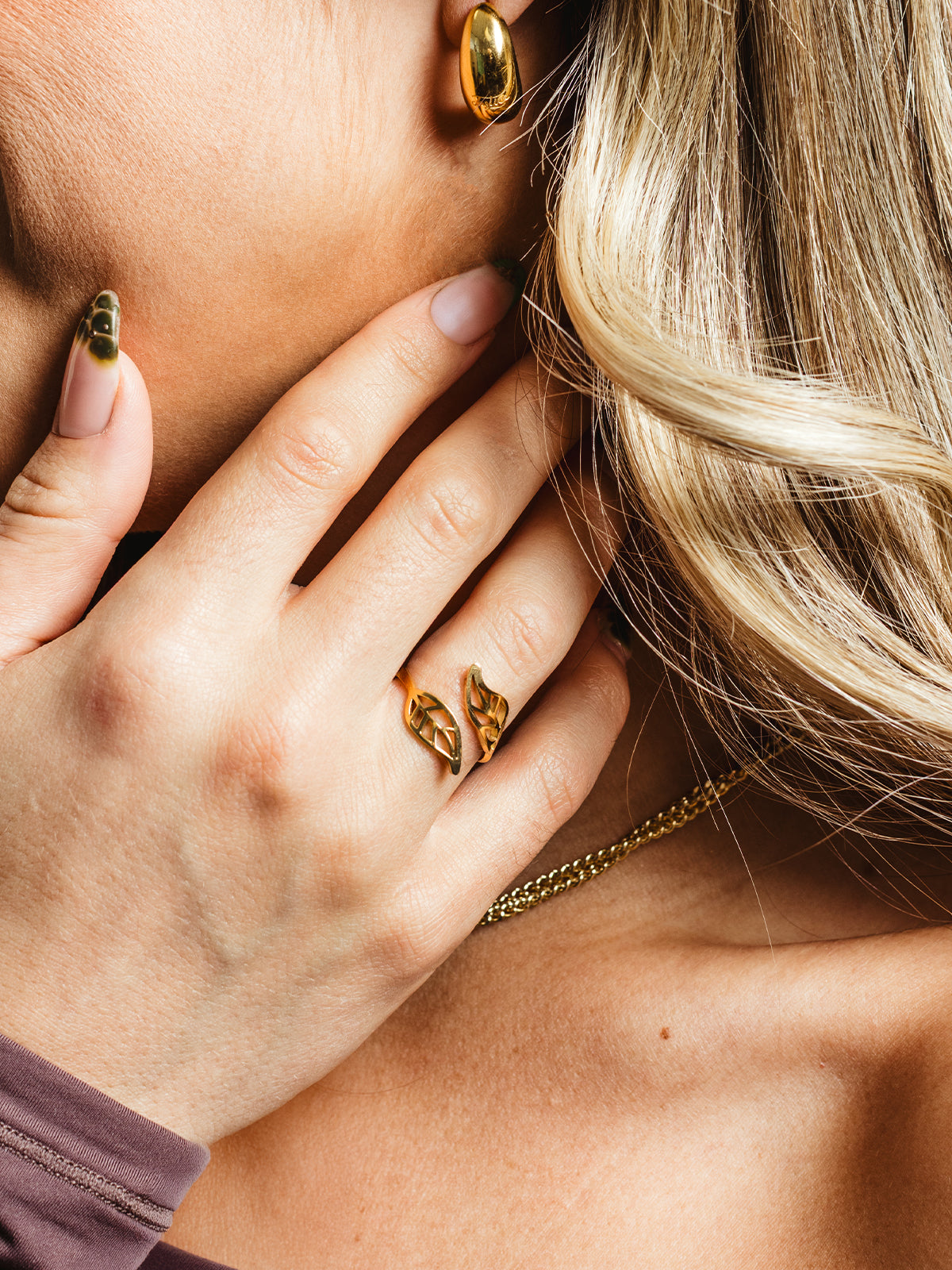 Gold leaf-shaped wrap ring worn on a woman's hand near her face, styled with a gold earring and blonde hair in soft waves.