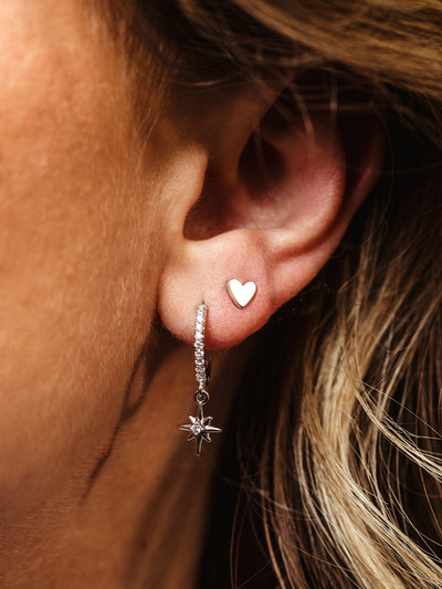 Close-up of a woman's ear wearing a small silver heart stud in the upper piercing and a pavé hoop earring with a dangling star charm in the lower piercing.