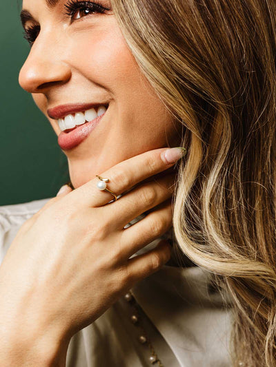 Close-up of a smiling woman wearing a gold ring with a single pearl accent, touching her chin. Her blonde hair frames her face and she wears a neutral-toned blouse, showcasing elegant handcrafted jewelry.