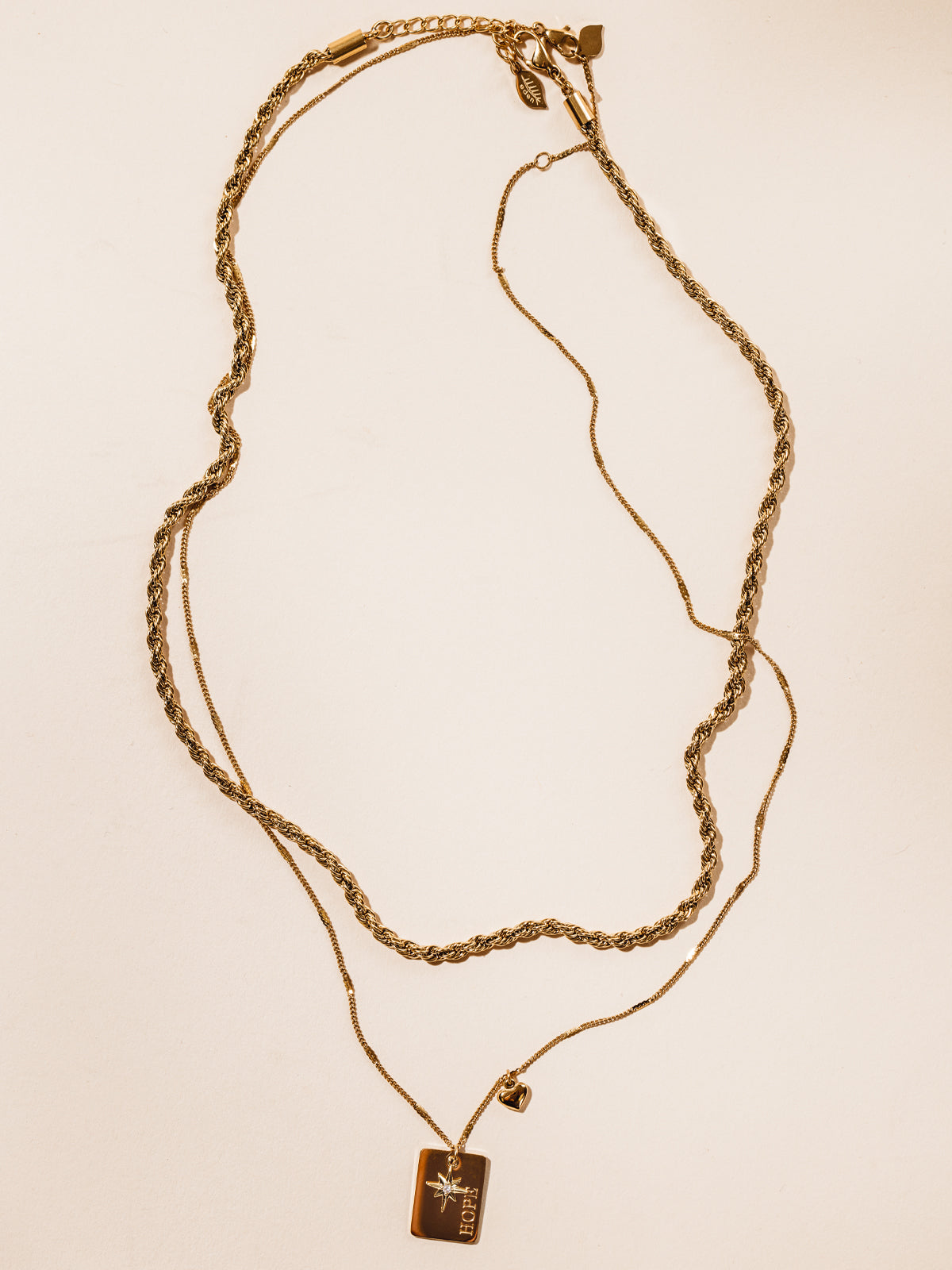 Statement gold necklace featuring two chains and rectangular pendant with "hope" etched into pendant. Also featuring a heat and star charm. Photographed on cream background. 