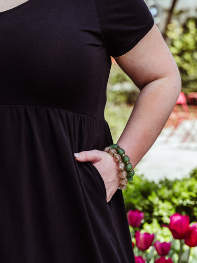 Close-up of a woman wearing a black short-sleeve dress with her hand in the pocket. She accessorizes with stacked green and neutral-toned beaded bracelets, standing in front of blooming tulips.