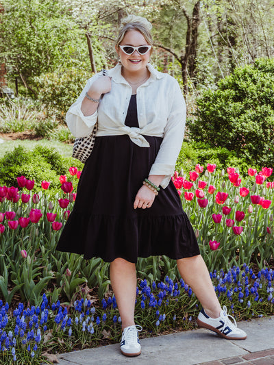 Woman stands smiling in a tulip garden wearing a black ruffle-hem midi dress layered with a tied white button-up shirt. She accessorizes with white cat-eye sunglasses, beaded bracelets, sneakers, and a checkered shoulder bag.