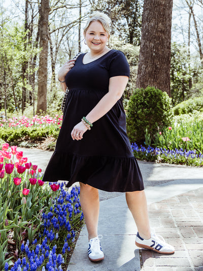 Woman in a black tiered midi dress poses in a sunny garden with colorful tulips. She wears casual sneakers, a checkered bag, green beaded bracelets, and minimal jewelry for a playful spring look.