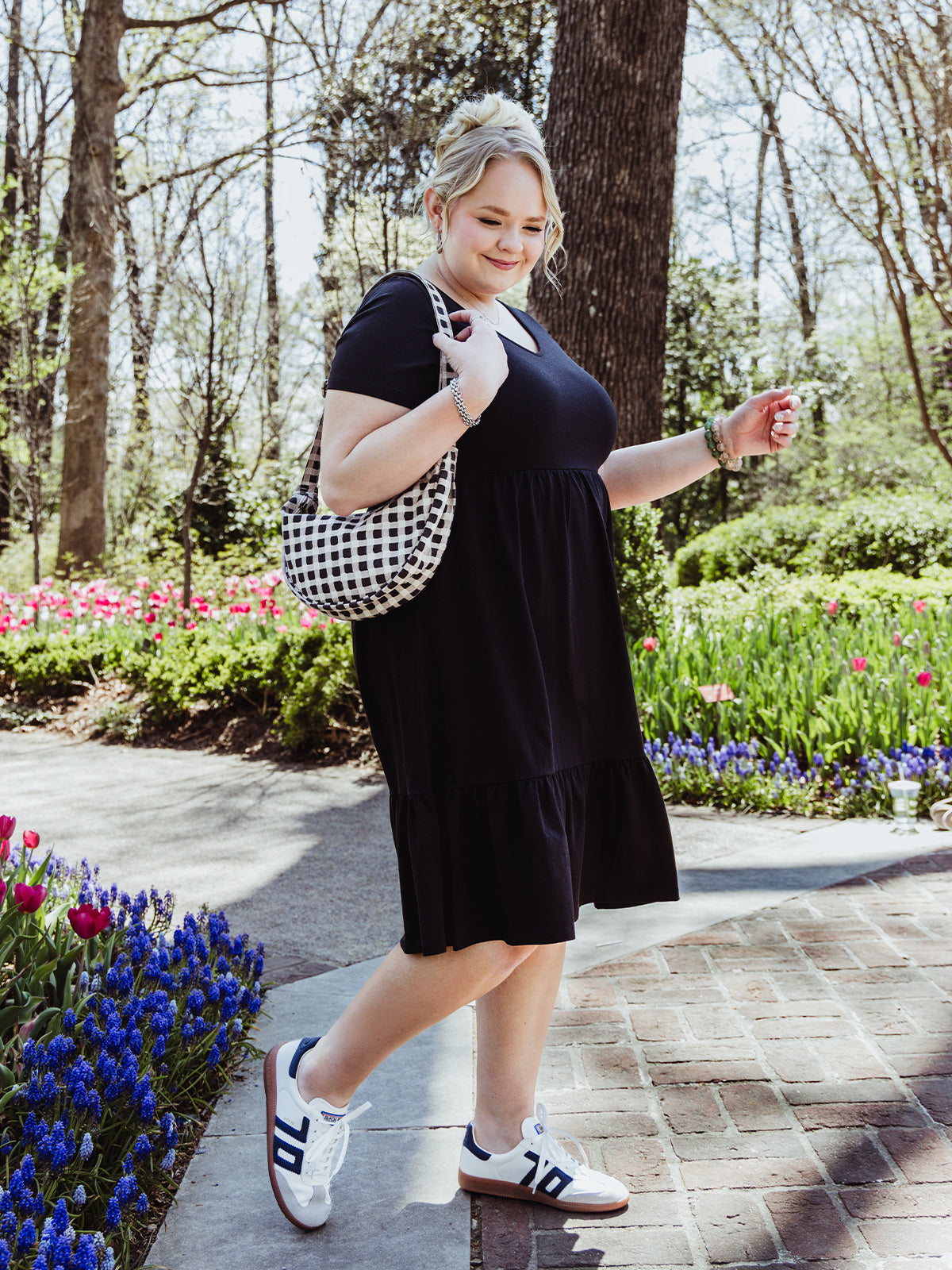 Side view of a woman walking confidently in a black short-sleeve midi dress and sneakers. She carries a black and white checkered shoulder bag and wears a green beaded bracelet, surrounded by vibrant spring blooms.