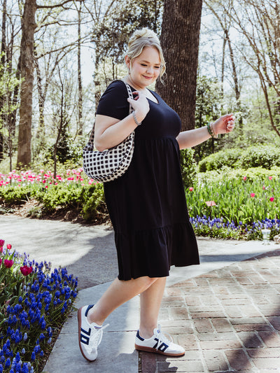 Side view of a woman walking confidently in a black short-sleeve midi dress and sneakers. She carries a black and white checkered shoulder bag and wears a green beaded bracelet, surrounded by vibrant spring blooms.