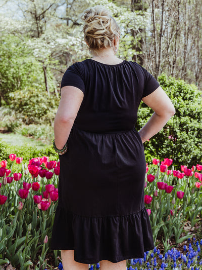Back view of a woman wearing a black midi dress with short sleeves and a gathered neckline. She stands among bright pink tulips, showing off her curly updo and green beaded bracelet.