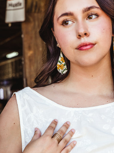 A young woman with long dark hair wears a white sleeveless top with delicate embroidery. She touches her collarbone with one hand, displaying a gold ring, while her beaded earrings in green, yellow, and white tones stand out against her dark hair. The background is slightly blurred, highlighting her thoughtful expression.