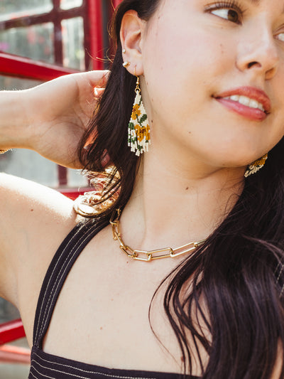 A young woman with long dark hair poses in front of a red window frame, wearing a black striped top. She adjusts her hair with one hand, showing off a pair of green, yellow, and white beaded earrings and a chunky gold chain necklace. The bright sunlight highlights her joyful expression and the intricate details of her jewelry.