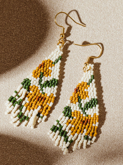 Close-up of beaded earrings with a sunflower pattern, using yellow, green, and white beads. The earrings are displayed on a sandy beige background, emphasizing their bold and intricate design.