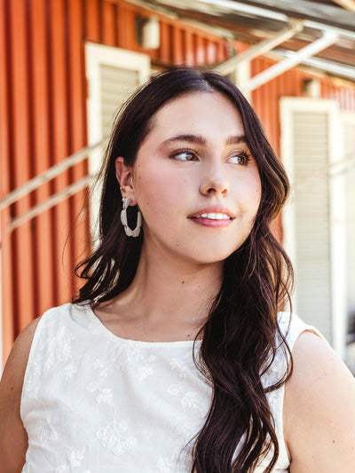 Woman wearing handcrafted white leather hoop earrings with a semi-circle design, styled with a white embroidered top, in an outdoor setting.