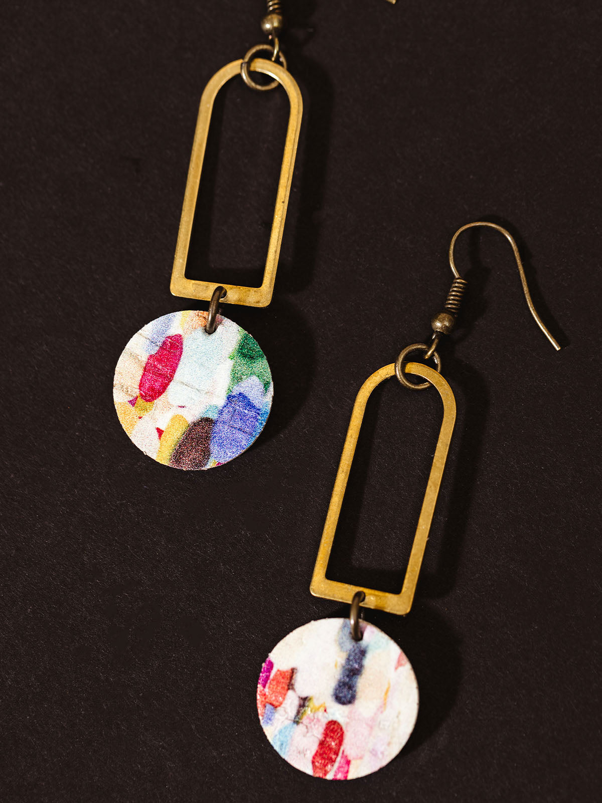 Close-up of handcrafted geometric earrings with a brass rectangular frame and a colorful circular pendant, displayed on a dark background.