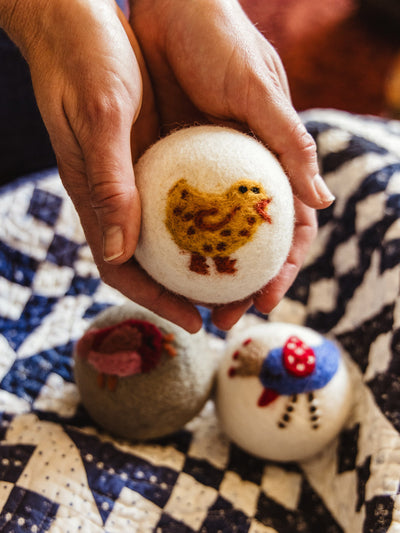 Hands holding a wool dryer ball featuring a yellow felted chicken, with two additional bird-themed dryer balls—one gray with a red bird and one white with a blue and red chicken—resting on a quilted blanket.