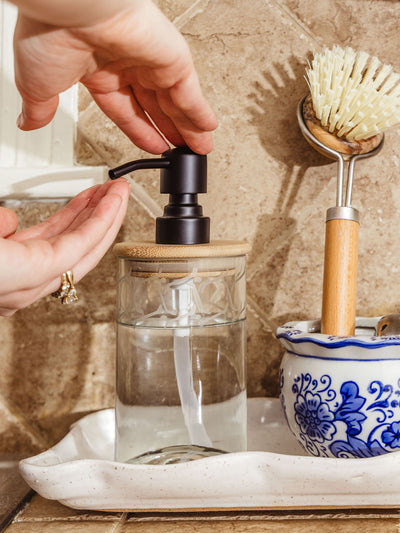 Close-up of hands using a glass soap dispenser with a black pump and bamboo lid, sitting on a ceramic tray next to a ceramic dish brush in a blue and white holder on a tiled kitchen counter.
