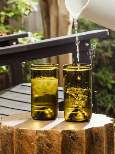Water is being poured from a white pitcher into one of two olive green glasses filled with ice, sitting on a wooden outdoor table with a black bench in the background.